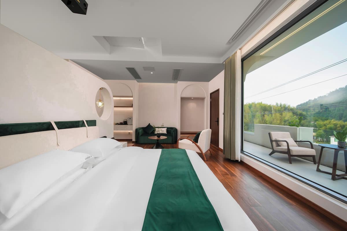 A hotel room with a large bed, a green runner, a seating area with a table and chairs, and a balcony with a view of the outdoors.
