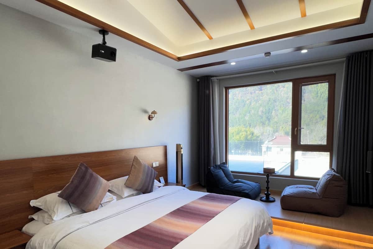 A modern hotel room with a large bed, wooden ceiling beams, a view of the outdoors, and a seating area with a table and chairs.