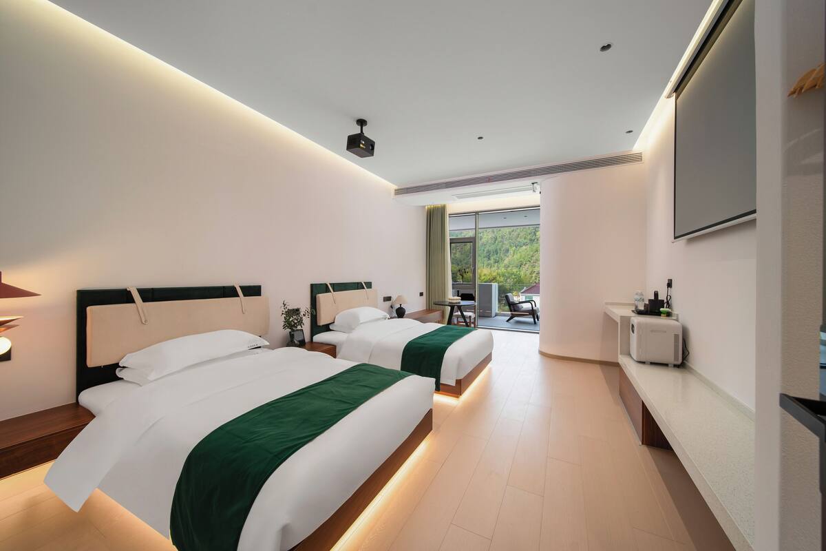 A hotel room with two beds, a desk, a chair, a TV, and a view of greenery outside.