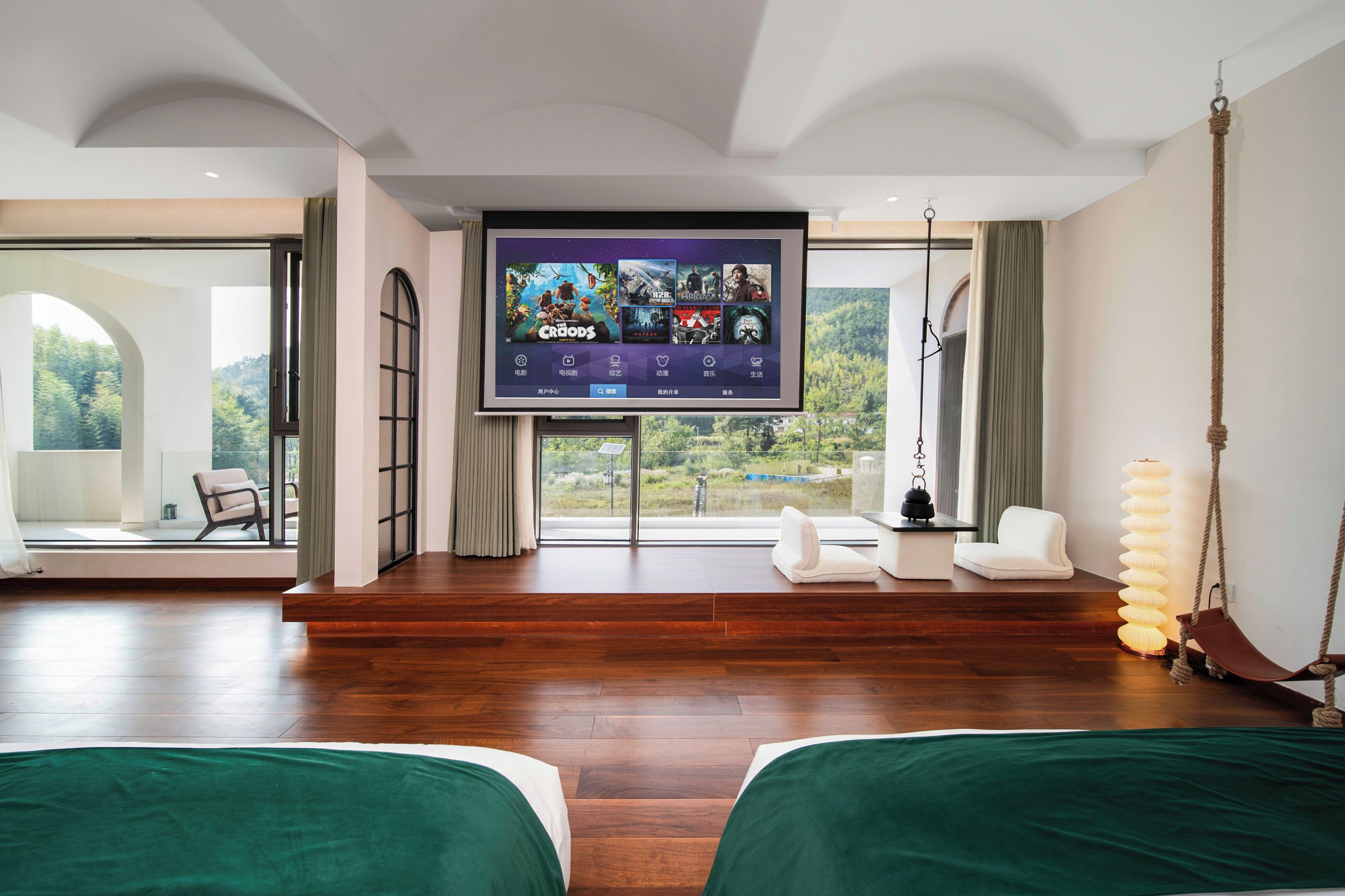 A hotel room with a large TV screen displaying game titles, a sofa, a chair, a hanging swing, and a view of the outdoors.