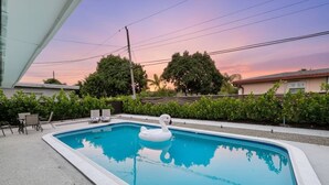 A rectangular swimming pool with a swan float, surrounded by a concrete deck, chairs, and a view of a sunset and residential buildings. - Modern King Suite Cozy Sofa Bed Near Everything (Pembroke Pines)
