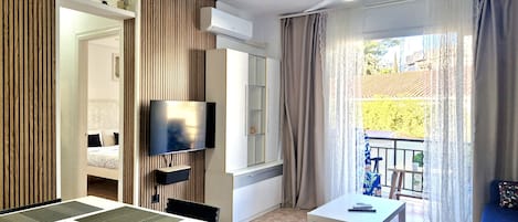 Moderni hotellihuone, jossa on ruokailutila, televisio, kattotuuletin ja parveke, jolta on näkymä.