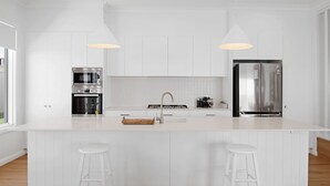 A modern kitchen with white cabinetry, a central island with stools, a stainless steel refrigerator, and a built-in oven. - The Whitehaus. Contemporary Calm. Wagga Wagga. (Gobbagombalin)