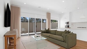 A modern living room with a glass coffee table, a green sofa, and a kitchen area with white cabinets and a microwave. - The Whitehaus. Contemporary Calm. Wagga Wagga. (Gobbagombalin)