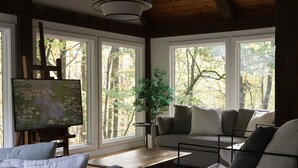 Uma sala de estar aconchegante com teto de madeira, uma pintura em um suporte, um sofá, uma mesa de centro e janelas amplas com vista para árvores.