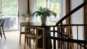 Um quarto espaçoso com vigas de madeira no teto, uma mesa redonda de jantar com cadeiras, um vaso de planta e uma vista através de grandes janelas.