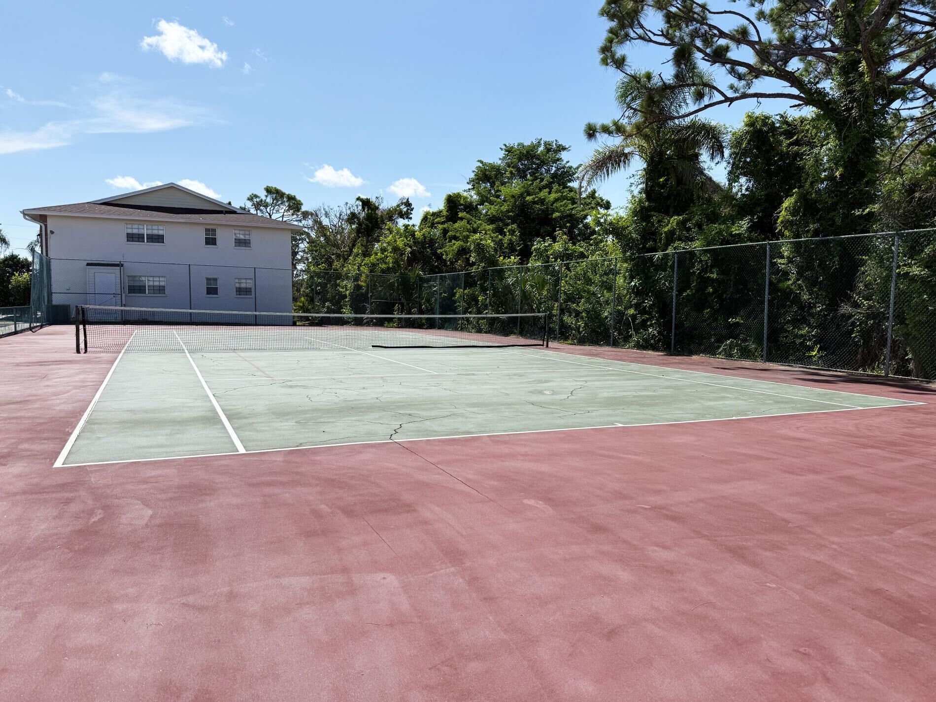 Una cancha de tenis exterior con una red, rodeada por una valla y árboles.
