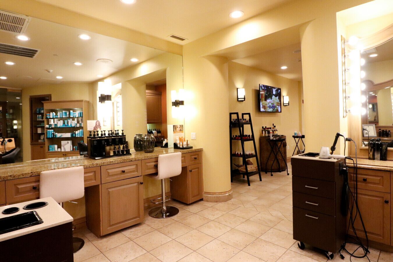 A well-lit salon with a large mirror, multiple chairs, and a vanity area with a sink and various beauty products.