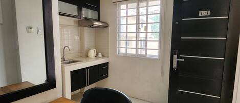 Una cocina compacta con una mesa de comedor de madera, una silla negra, una ventana con persianas y una puerta negra con el número 101.