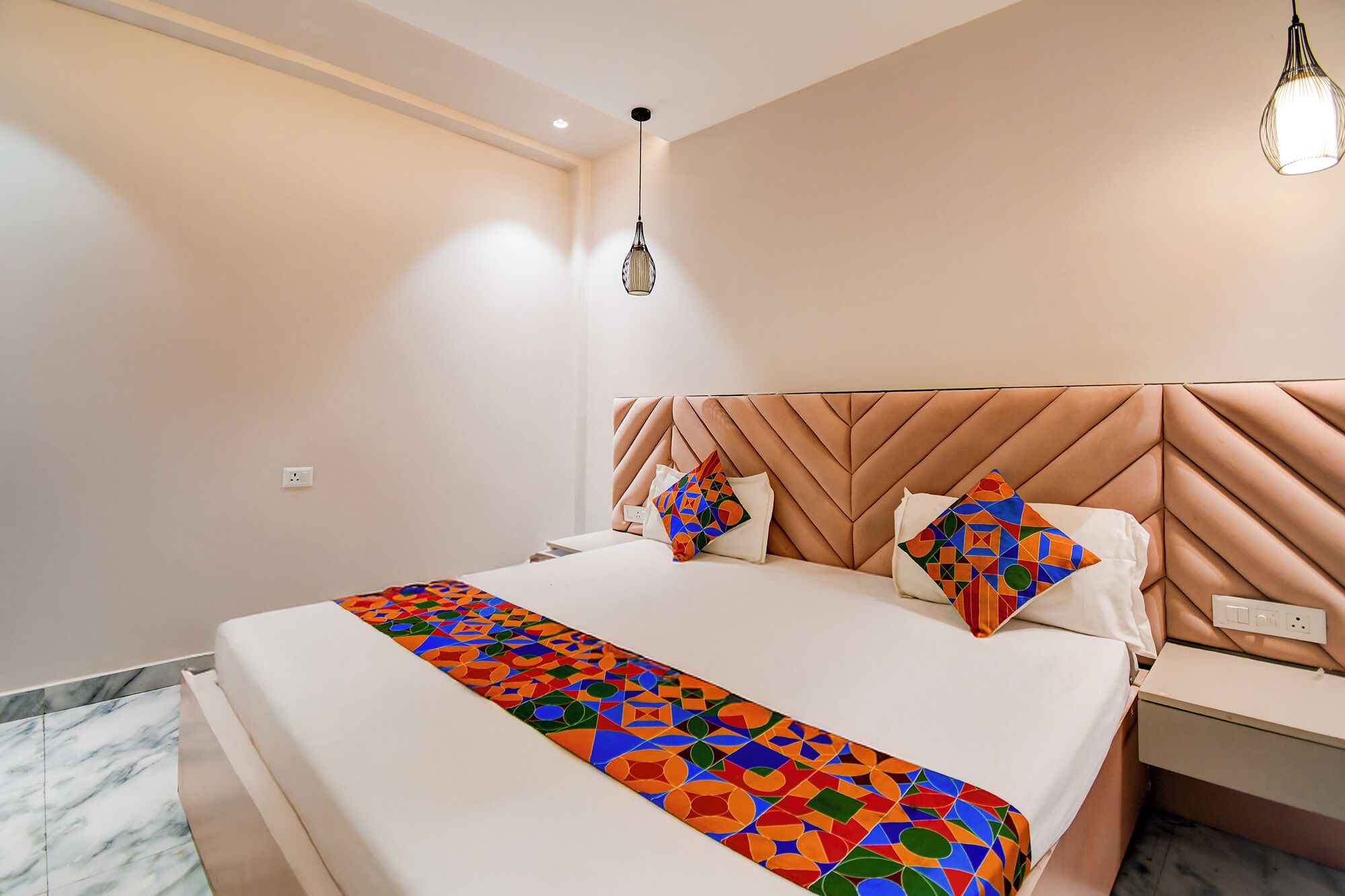 A modern hotel room with a large bed, a geometric-patterned bedspread, a tufted headboard, and a nightstand with a lamp.