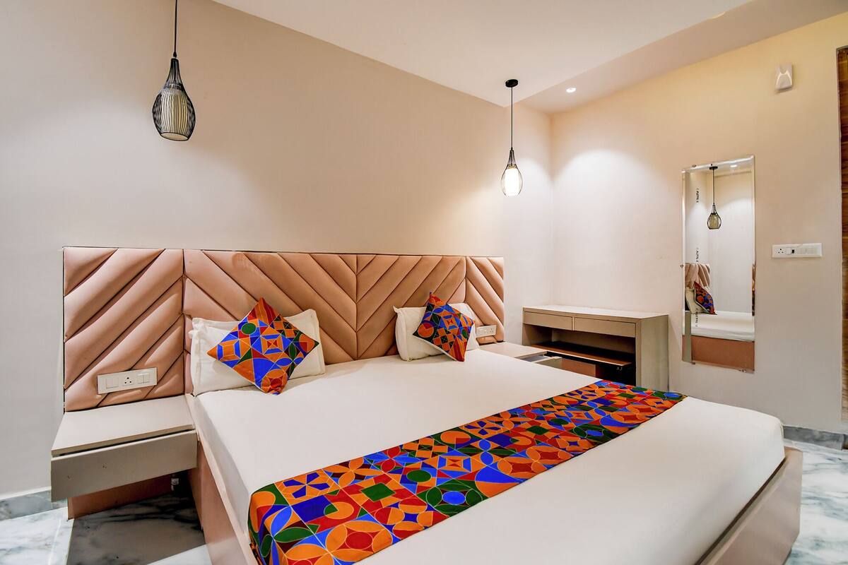 A modern hotel room with a large bed, a headboard with geometric patterns, a bedside table, a nightstand, a mirror, and a hanging light fixture.