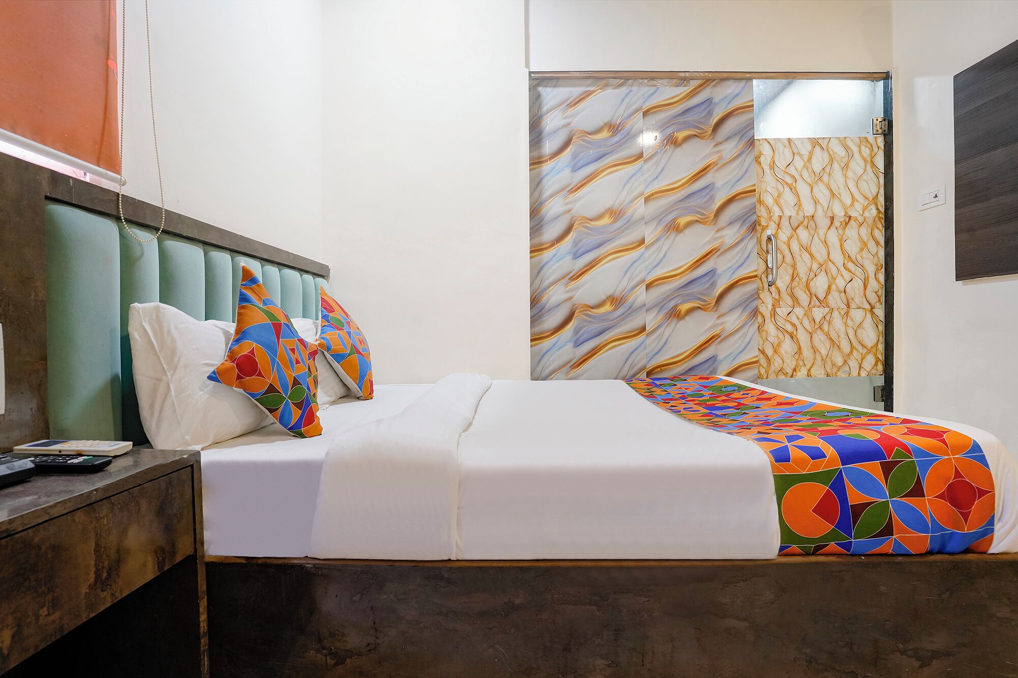 A neatly made bed with a colorful geometric-patterned blanket, a wooden nightstand with a remote control, and a headboard with a unique design.