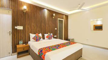 Quarto de hotel com parede de painel de madeira, cama com roupa de cama colorida, mesa de cabeceira com uma garrafa, criado-mudo e um ventilador de teto, além de um espelho na parede.