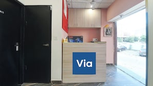 A reception desk with a 'Via' sign, a black door, a ceiling fan, a red flag, and a framed picture on the wall. - Via Capitol Square (Jaipur)