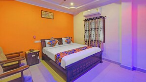 A bedroom with an orange wall, a bed with a colorful bedspread, a wooden nightstand, a chair, a ceiling fan, and an air conditioning unit. - Fabhotel Sri Balaji Grand (Hyderabad)