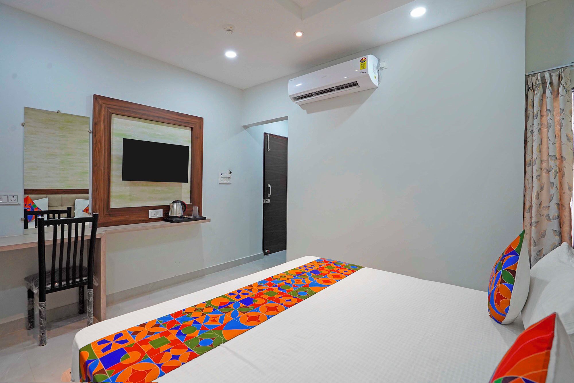 A hotel room with a bed, a desk with a chair, an air conditioning unit, and a door.