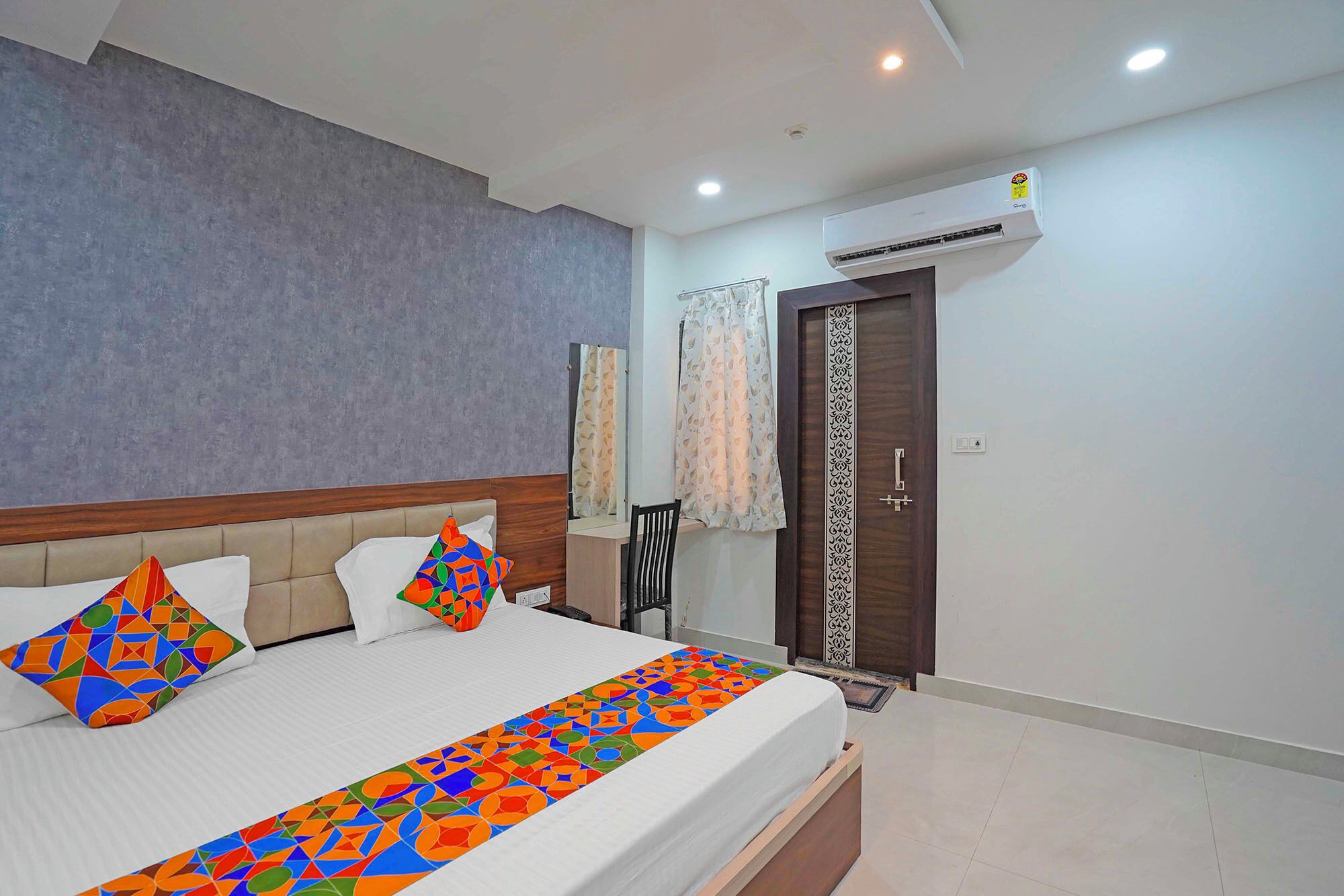 Une chambre d’hôtel comprenant un lit, une tête de lit en bois, une couvre-lit à motifs, une fenêtre avec des rideaux, un appareil de climatisation et une porte ornée d’un panneau décoratif.