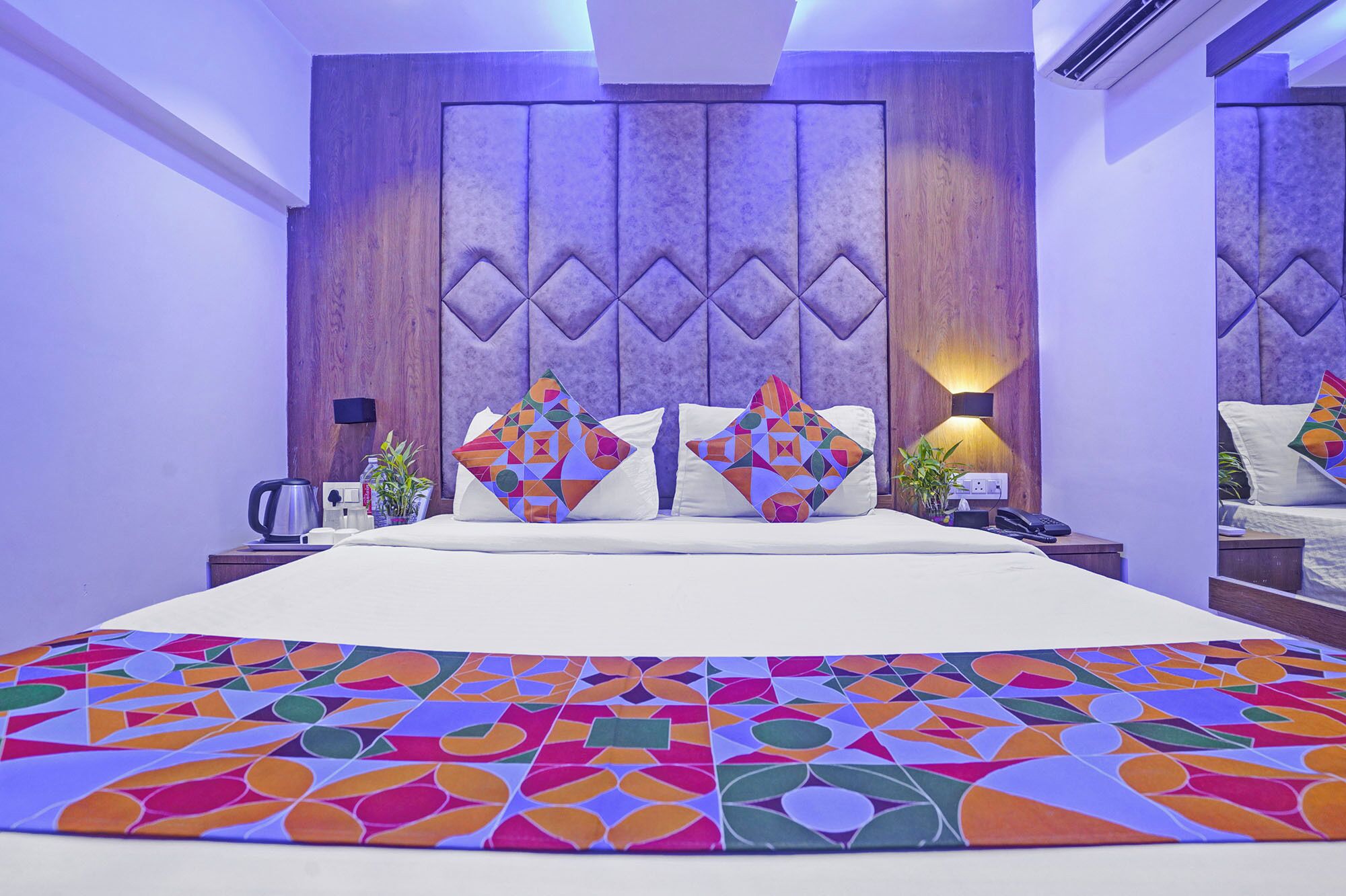 Quarto de hotel moderno com uma cama grande, almofadas com padrão geométrico, cabeceira de madeira e mesa de cabeceira com luminária e telefone.