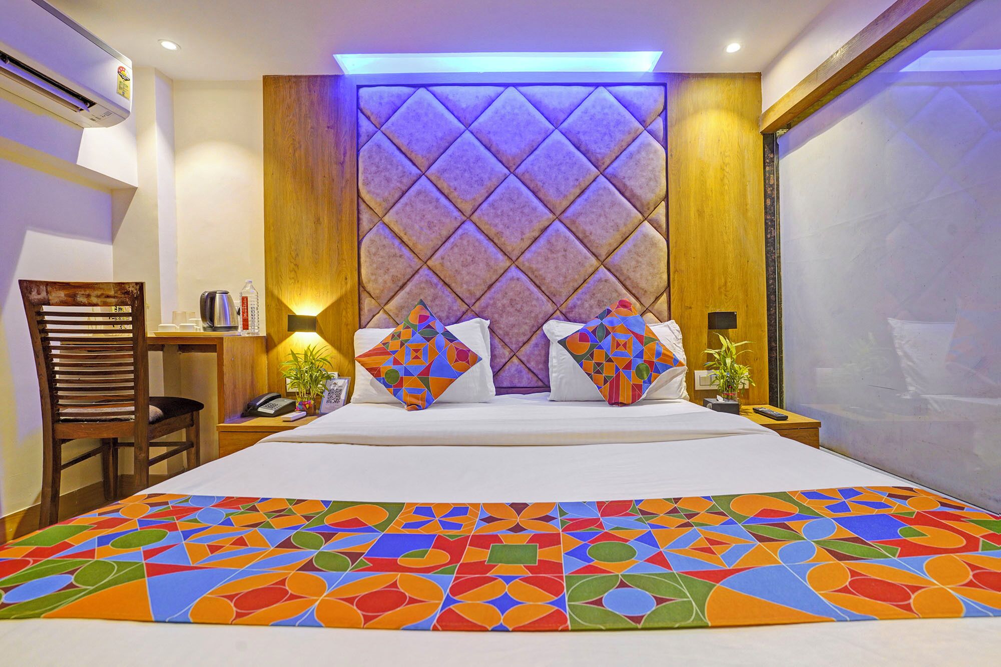 A hotel room with a large bed, a wooden headboard, a bedside table with a lamp, a chair, and a view of the outside.