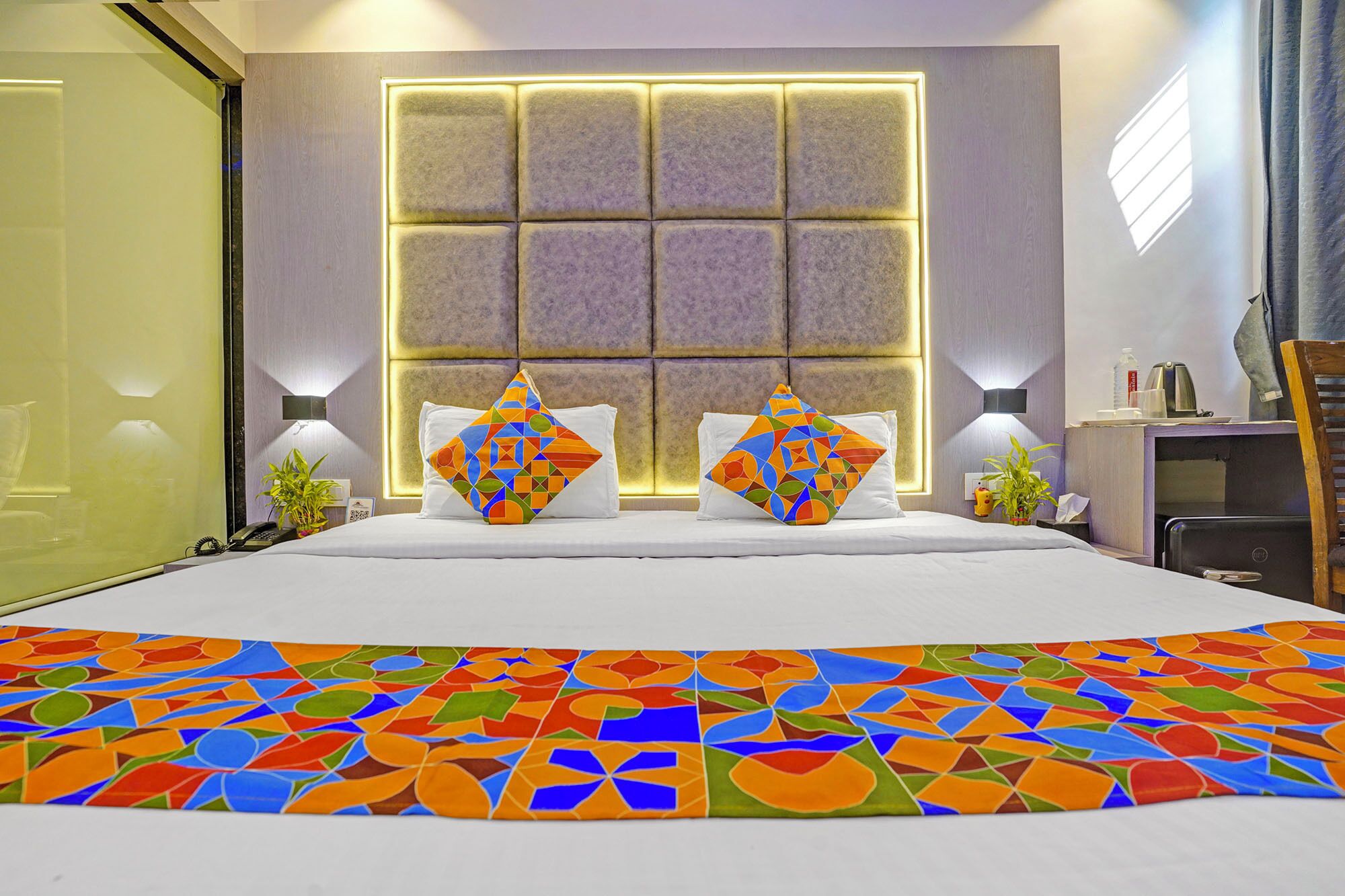 A hotel room with a large bed, two decorative pillows, a bedside table with a lamp, and a window with a light panel.