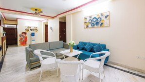 A living room with a blue sofa, white wicker chairs, a small table with a vase, and a ceiling fan. - Fabhotel Uk House Residency (Gurugram)