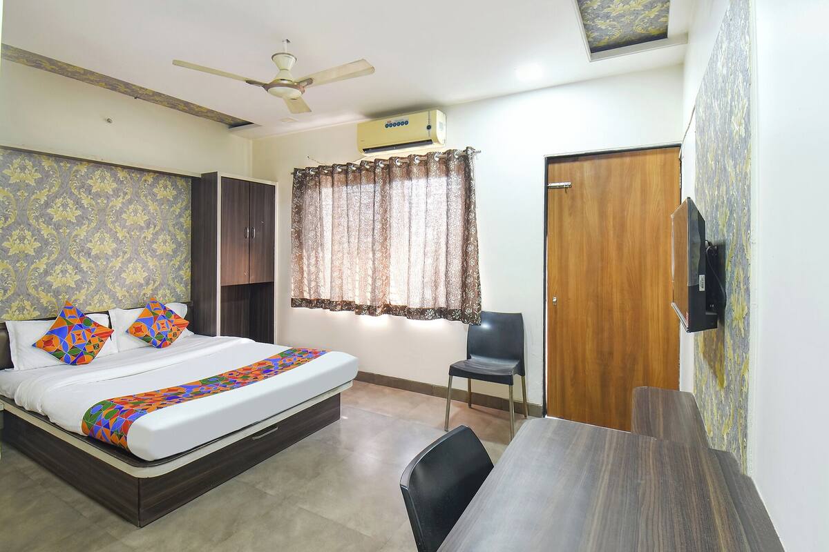 A hotel room with a bed, a desk, a chair, a wardrobe, a ceiling fan, an air conditioning unit, and a window with curtains.