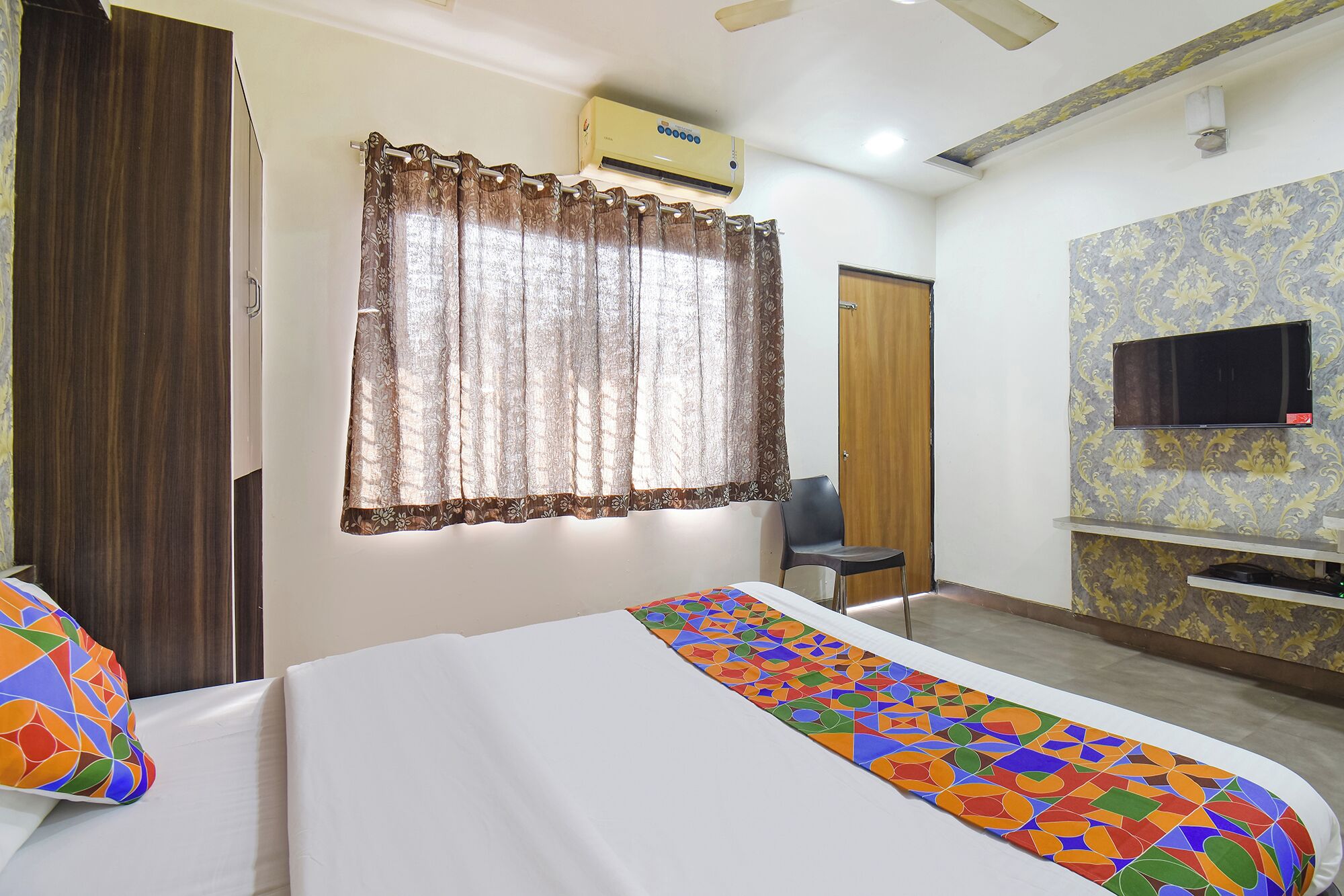A hotel room with a bed, a chair, a TV mounted on the wall, and an air conditioning unit.
