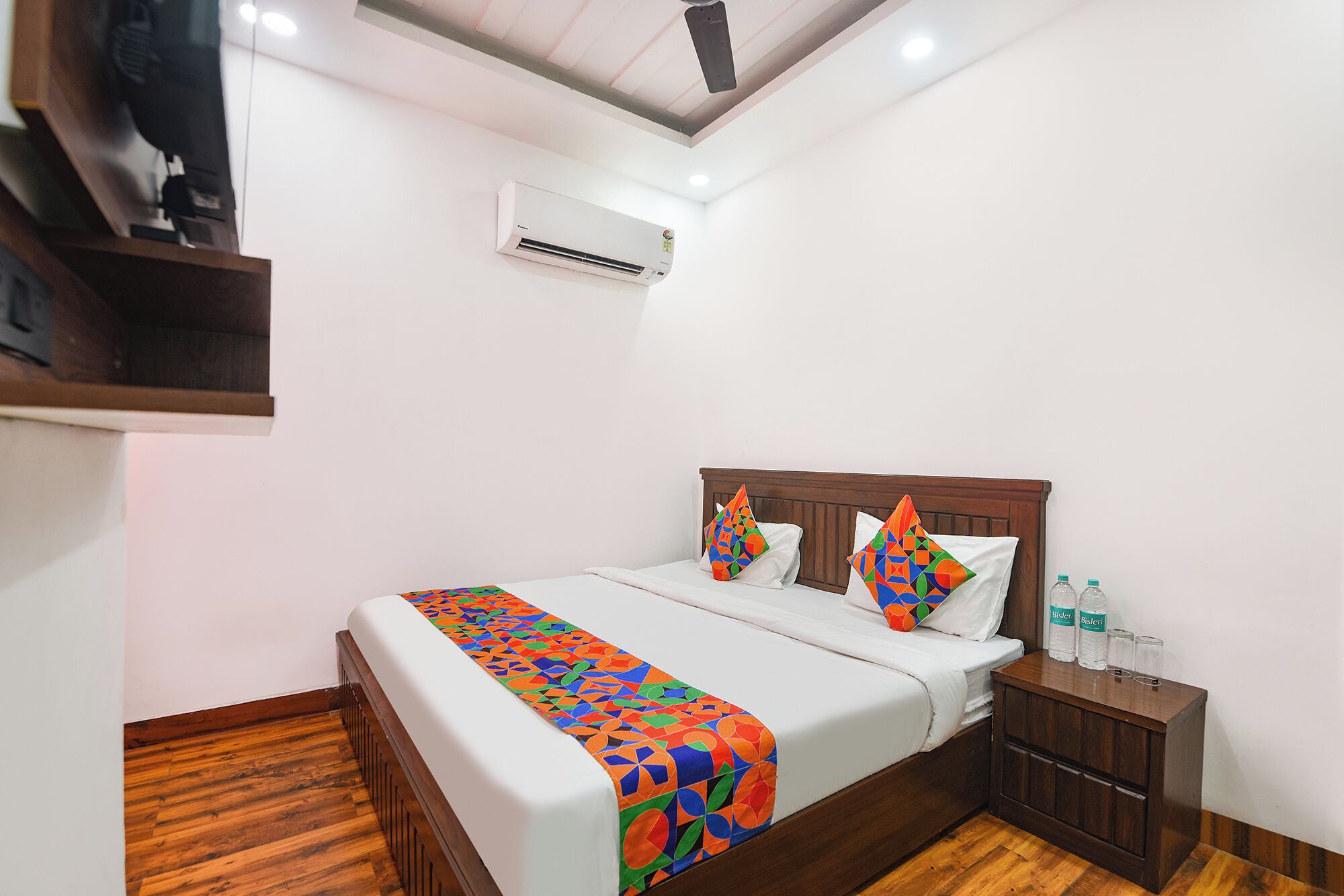 A hotel room with a wooden bed, colorful pillows, a white bedspread, a small wooden nightstand with bottles, and an air conditioning unit on the wall.