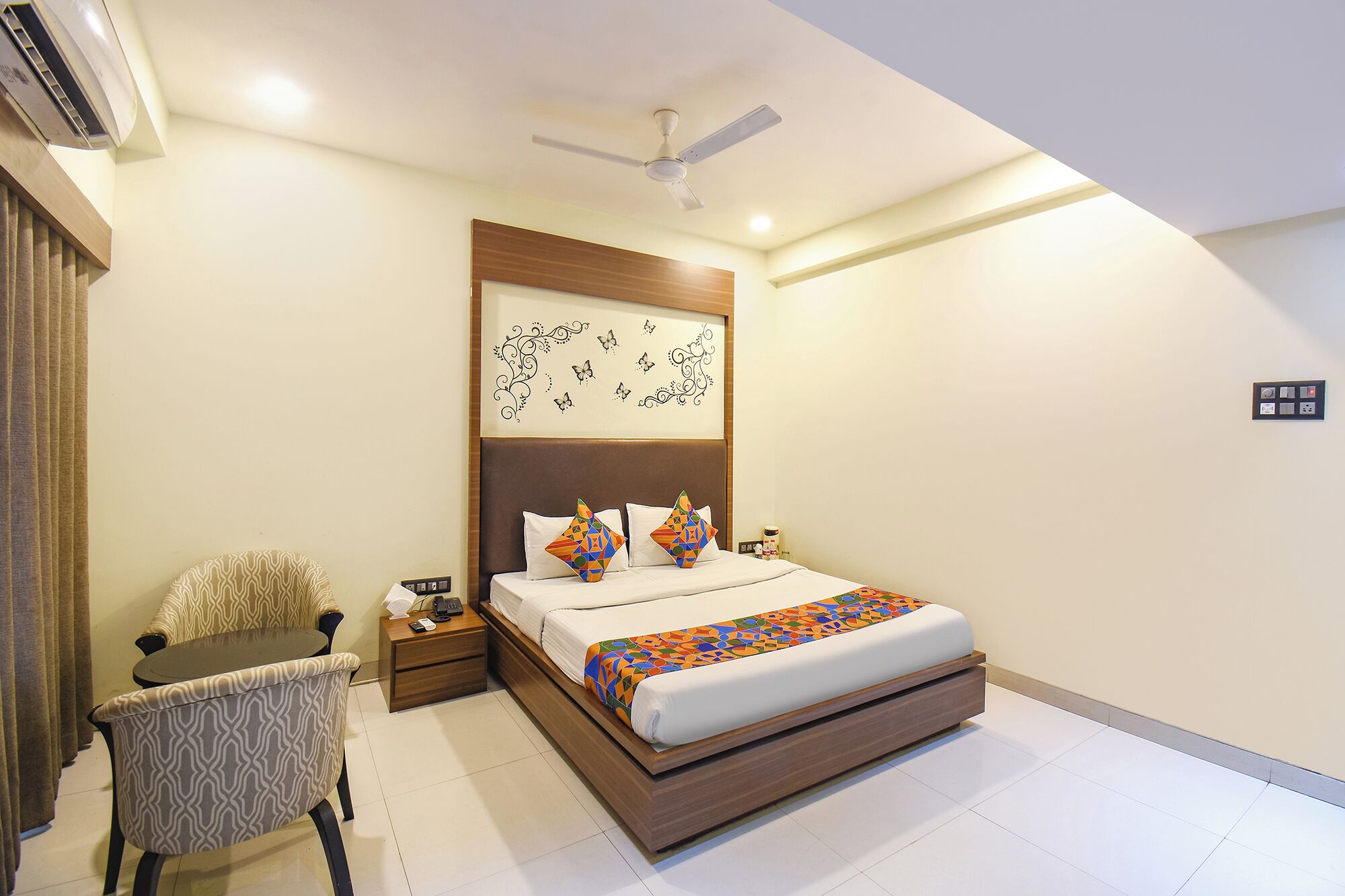 A modern hotel room with a bed, a chair, a nightstand, an air conditioning unit, a ceiling fan, and a decorative wall piece.