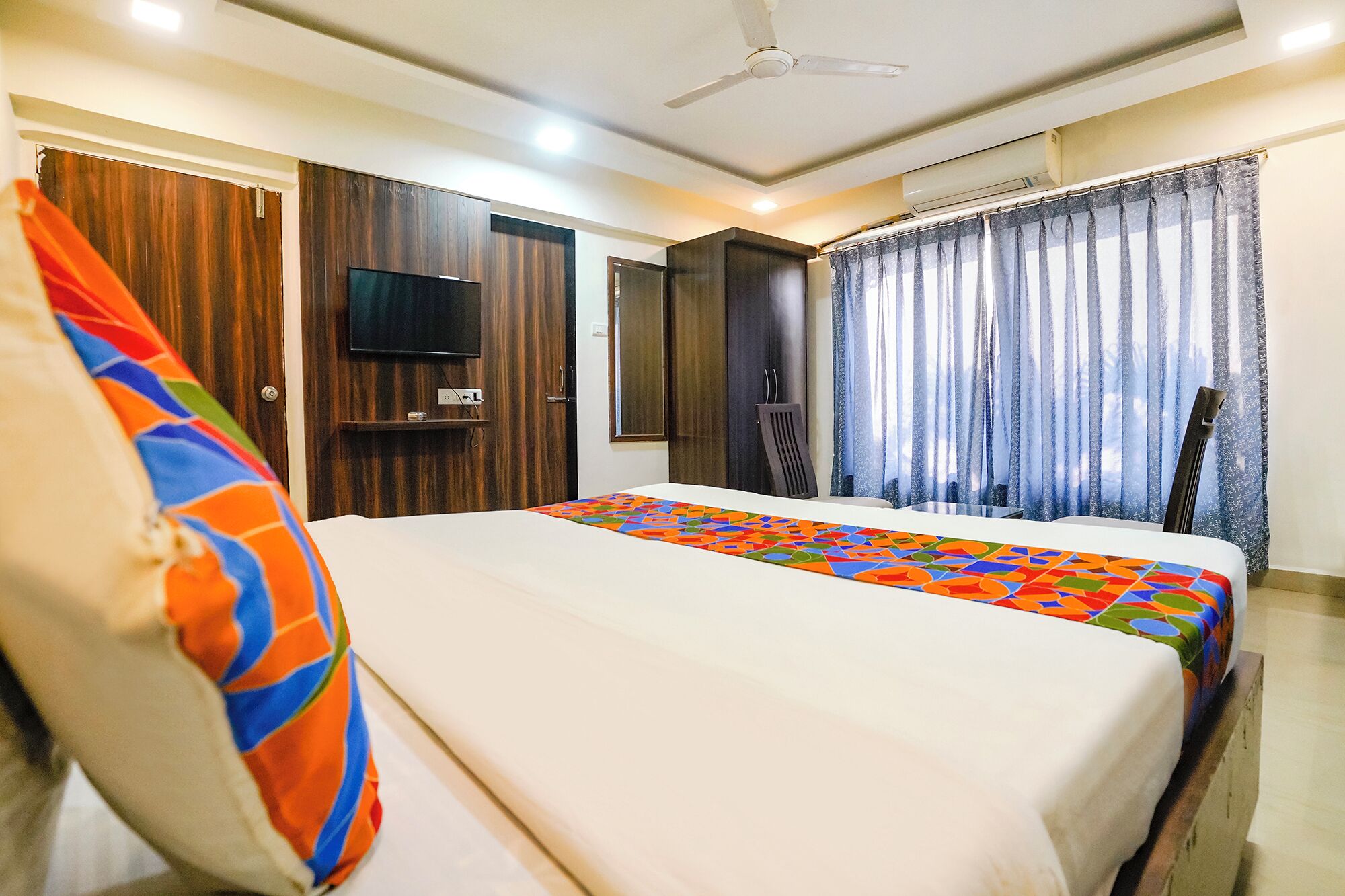 A hotel room with a bed, a TV mounted on the wall, a ceiling fan, and a window with curtains.