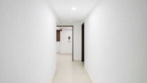 Un corridoio stretto e ben illuminato, con pareti bianche e pavimento piastrellato, che conduce a una stanza con una porta e una piccola finestra.
