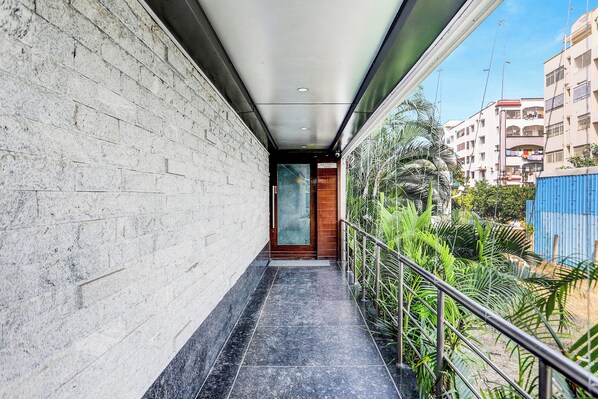 Un couloir étroit, doté d’un sol carrelé, d’une porte en bois et offrant une vue sur un quartier résidentiel avec ses immeubles et sa verdure.