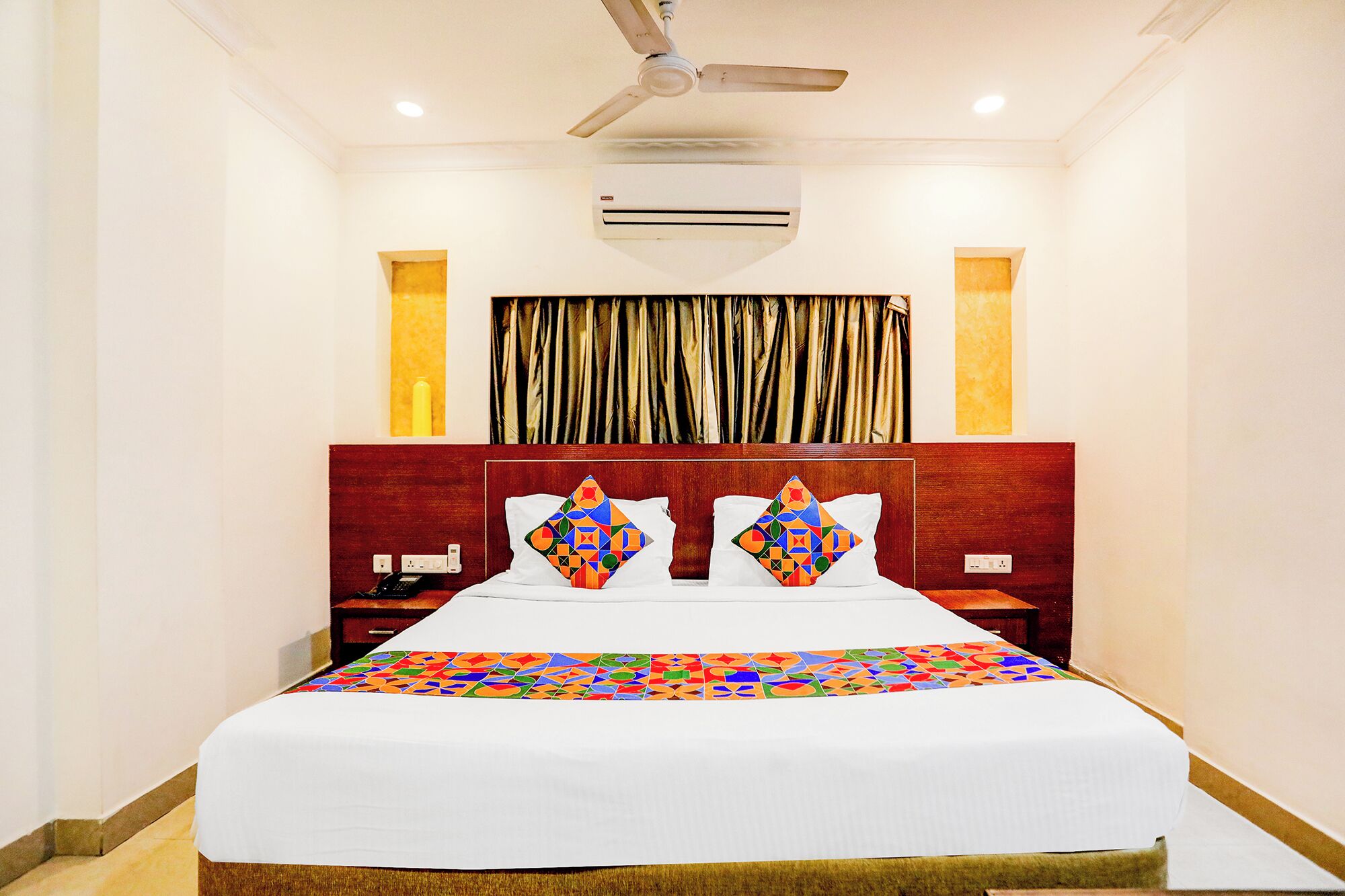 A hotel room with a bed, a ceiling fan, an air conditioning unit, and a window with curtains.