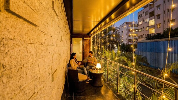 Un balcon agrémenté de meubles en osier, une table avec une lampe et une vue sur les bâtiments et la verdure.