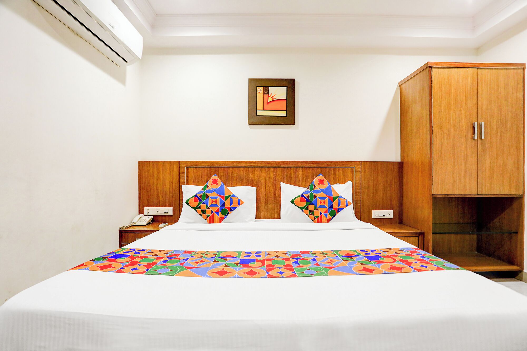 A hotel room with a bed, two bedside tables, a nightstand, a wooden wardrobe, and a wall-mounted artwork.