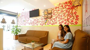 Un espace d’accueil avec un canapé marron, une table basse en verre et une fresque murale représentant un arbre et des fleurs.