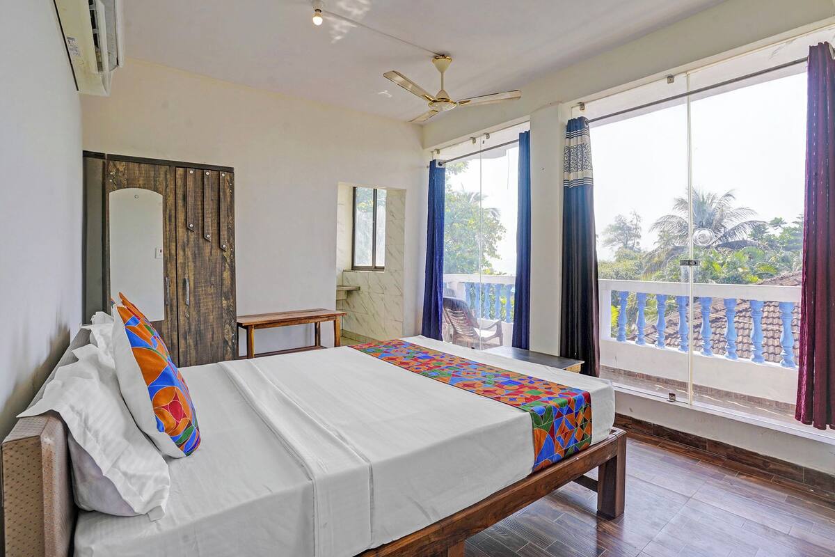 A hotel room with a bed, a wooden bench, a small table, a mirror, and a balcony with a view of palm trees.