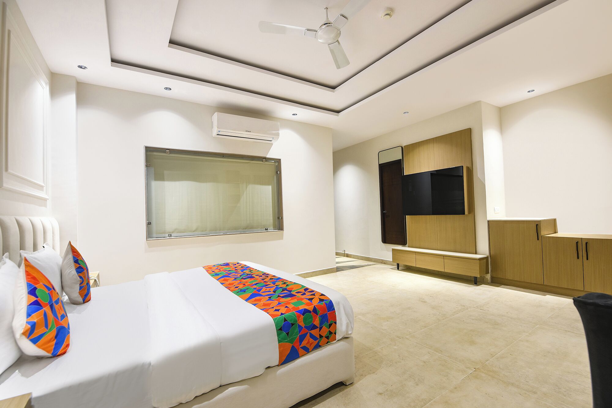 Une chambre d’hôtel moderne équipée d’un lit, d’une grande fenêtre, d’un ventilateur de plafond et d’un téléviseur fixé au mur.