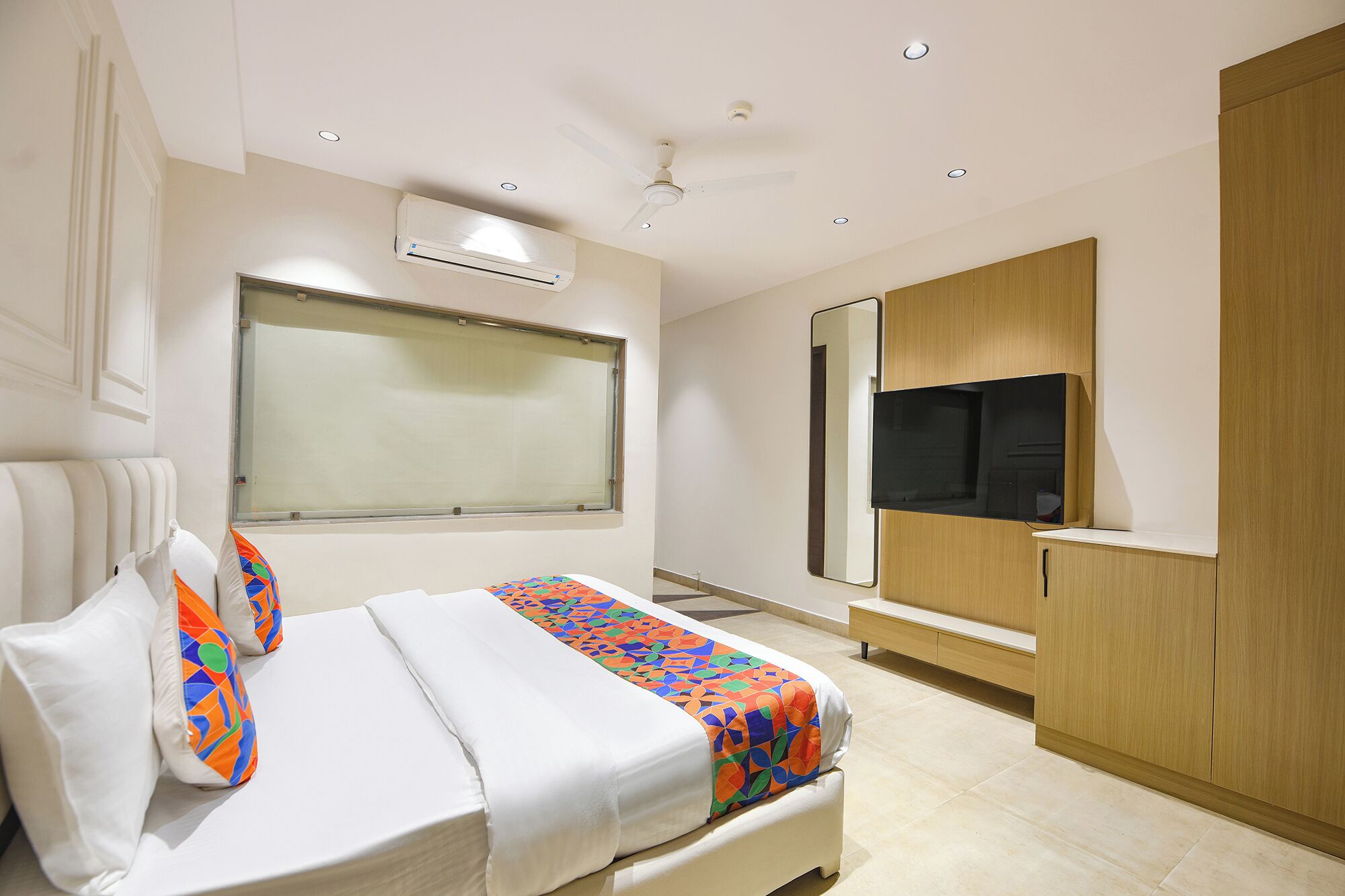 A hotel room with a bed, a large window, an air conditioning unit, a ceiling fan, a TV, and a wooden wardrobe.