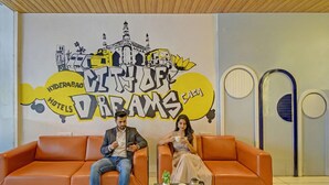 Two people sitting on an orange sofa in a hotel lobby with a mural of Hyderabad landmarks and the text "Hyderabad Hotels Dream House". - Fabhotel Casa Grand Himayathnagar (Hyderabad)