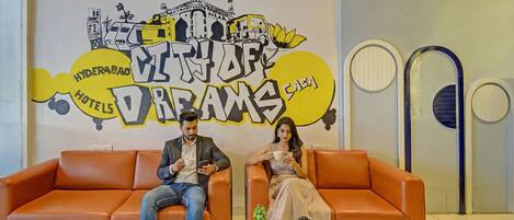 Two people sitting on an orange sofa in a hotel lobby with a mural of Hyderabad landmarks and the text "Hyderabad Hotels Dream House".