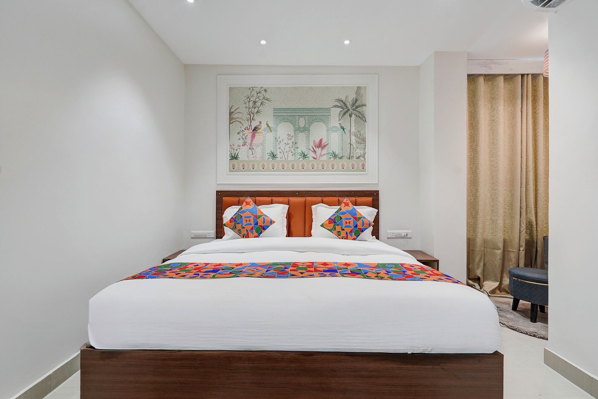 A hotel room with a large bed, a wooden headboard, a colorful geometric bedspread, and a framed artwork above the bed.