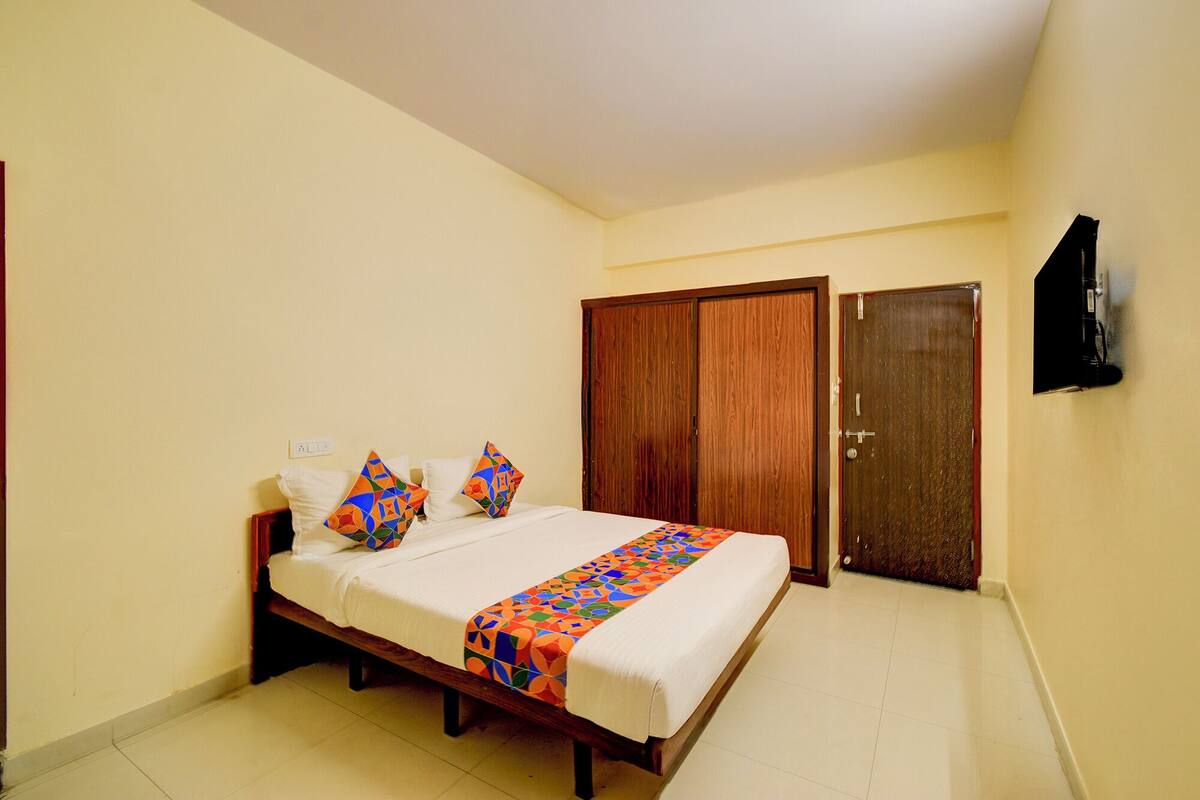 A bedroom with a bed, two pillows, a colorful blanket, a wooden wardrobe, and a television on the wall.