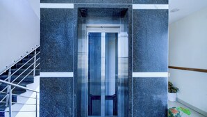 Ein moderner Aufzug mit Glastür, umrahmt von dunklen Steinwänden und einer Metalltreppe links.