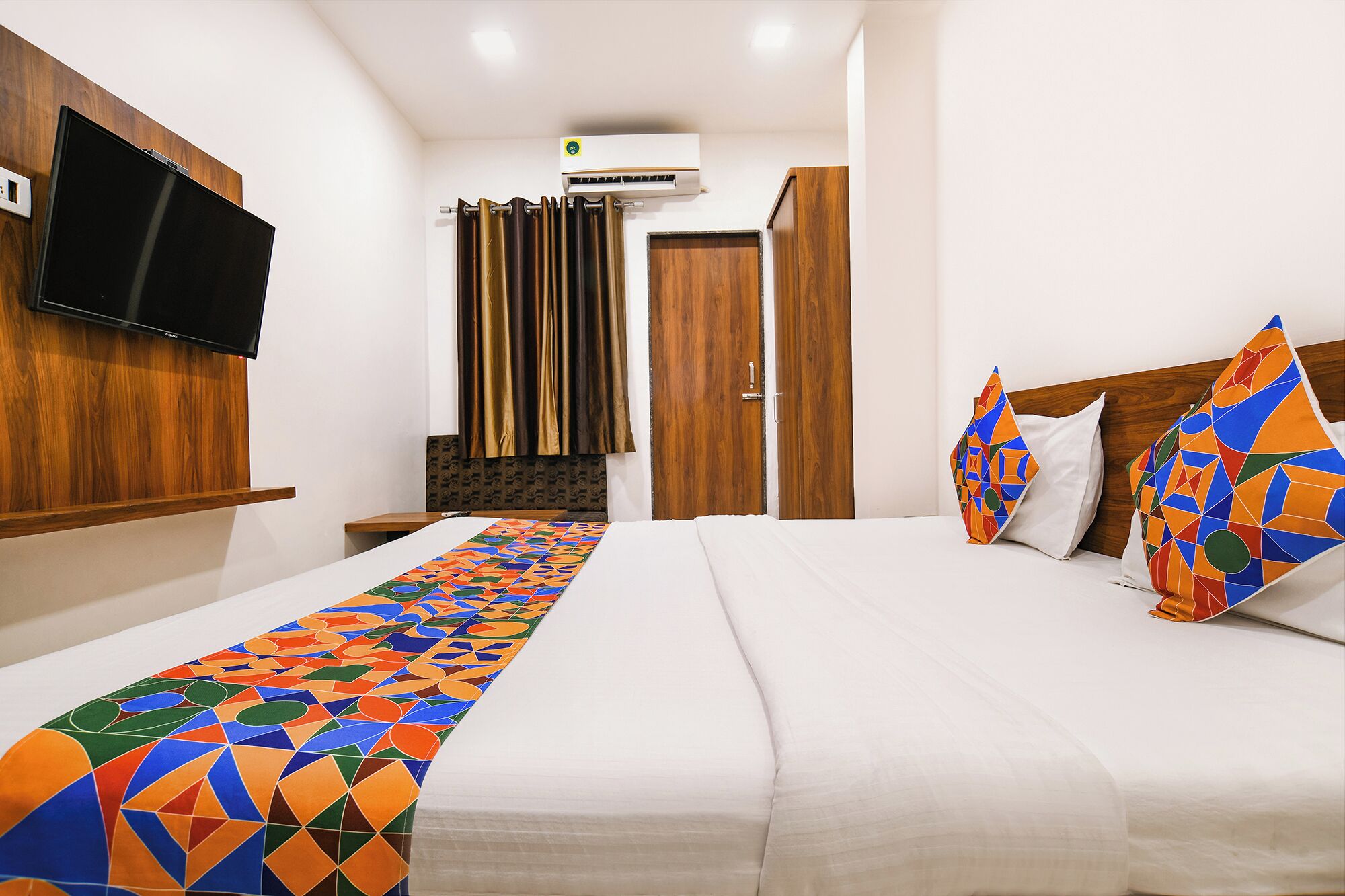 A hotel room with a bed, a TV mounted on the wall, a wooden wardrobe, and a curtain.