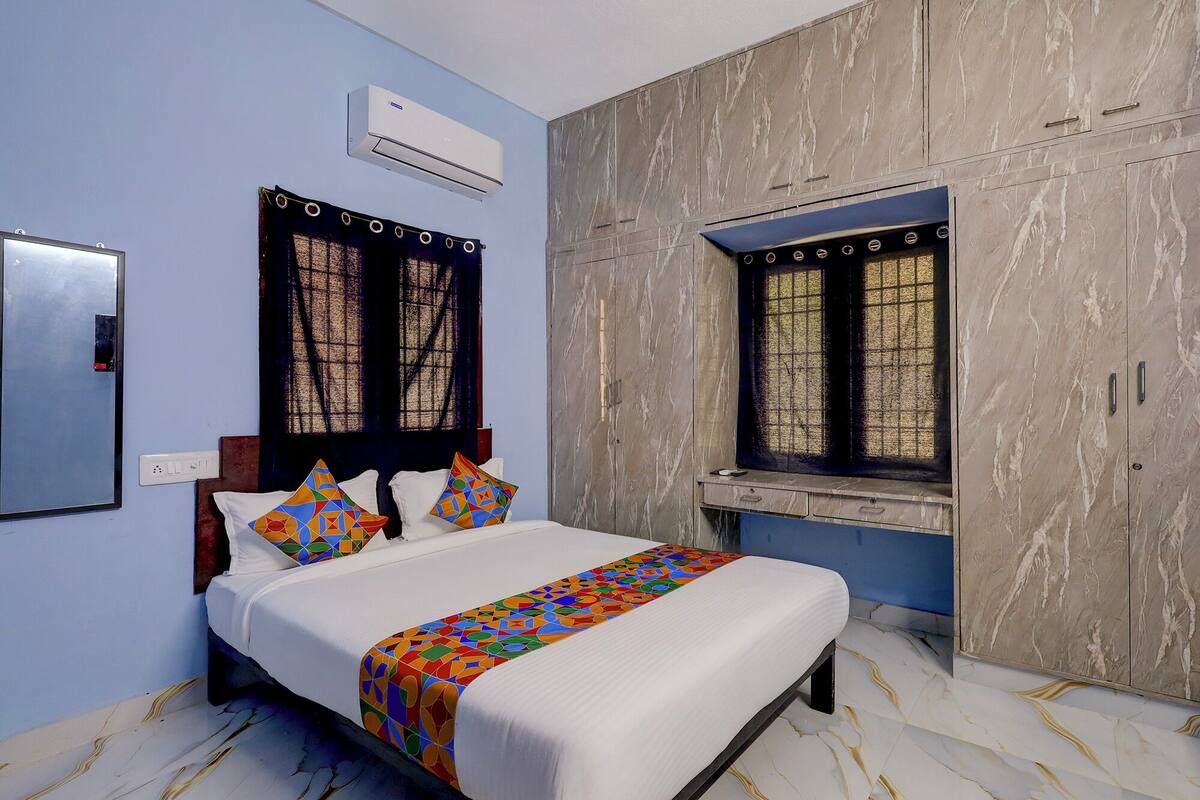 A bedroom with a bed, two windows with curtains, a wardrobe, and an air conditioning unit.