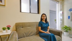 A woman sitting on a beige sofa in a room with a white wall, a framed picture, and a potted plant. - Fabhotel Stay Land Plus (Mumbai)