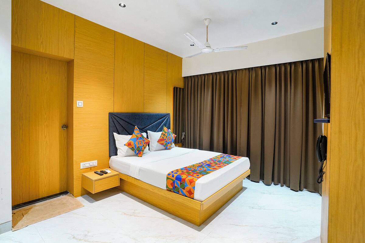 Une chambre d’hôtel moderne avec une tête de lit en bois, un lit agrémenté d’oreillers aux motifs géométriques et aux couleurs vives, une table de chevet avec un téléphone et une télécommande, et un ventilateur de plafond.