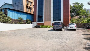A parking area with two cars parked in front of a modern building with vertical columns and a mix of glass and brick facade. - Fabhotel Gavalidev (Navi Mumbai)