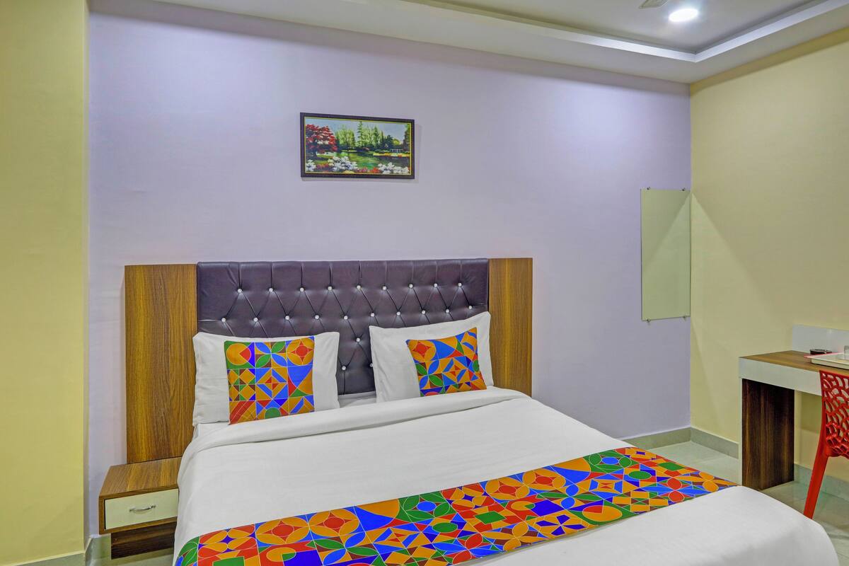 A bedroom with a tufted headboard, a bed with colorful geometric-patterned pillows, a nightstand, a desk with a chair, and a wall-mounted picture.
